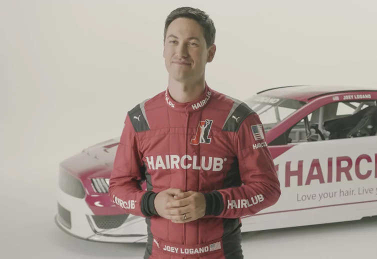 Celebrity Joey Logano - talking about his HairClub Xtrands+ solution for a commercial, standing in a white studio, wearing his red HairClub-sponsored racing suit with his HairClub racecar behind him.
