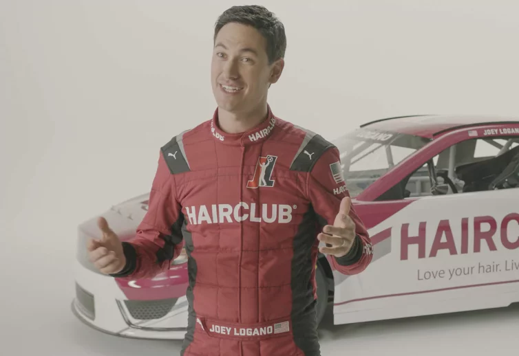 Celebrity Joey Logano - talking about his HairClub Xtrands+ solution, standing in a white studio, wearing his red HairClub-sponsored racing suit with his HairClub racecar behind him.