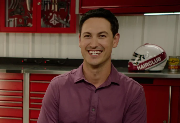 Celebrity Joey Logano - wearing a purple dress shirt, sitting in his HairClub racing team garage with a full head of hair because of his HairClub Xtrands+ solution.