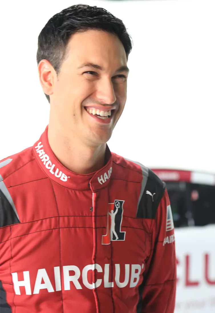 Celebrity Joey Logano - with a big smile, wearing his red HairClub-sponsored racing suit with his HairClub racecar behind him.