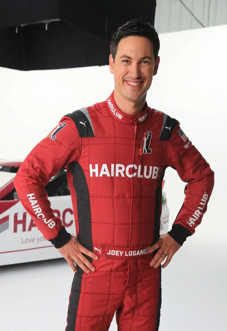 Celebrity Joey Logano - posing in his red HairClub-sponsored racing suit with his HairClub racecar behind him.