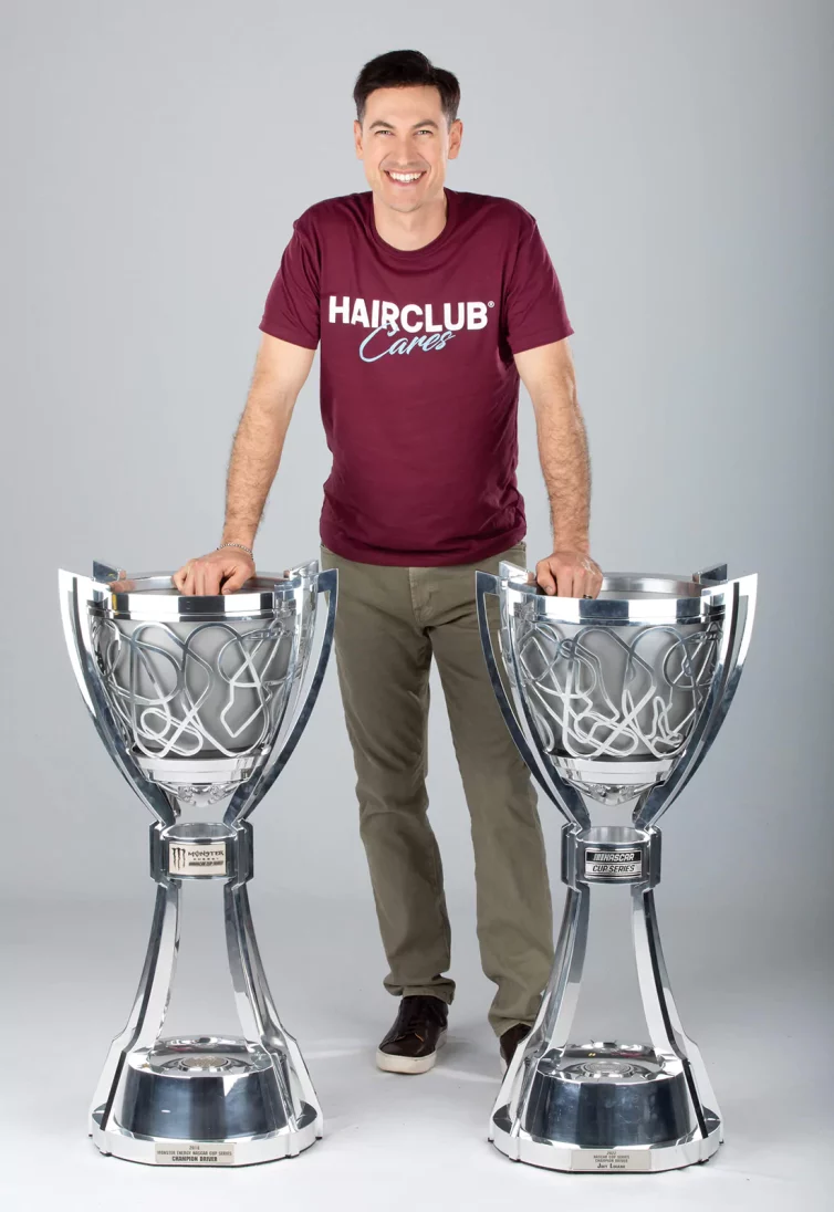 Celebrity Joey Logano - wearing a burgundy HairClub Cares t-shirt, standing between his 2 NASCAR Cup trophies with a hand on each one.