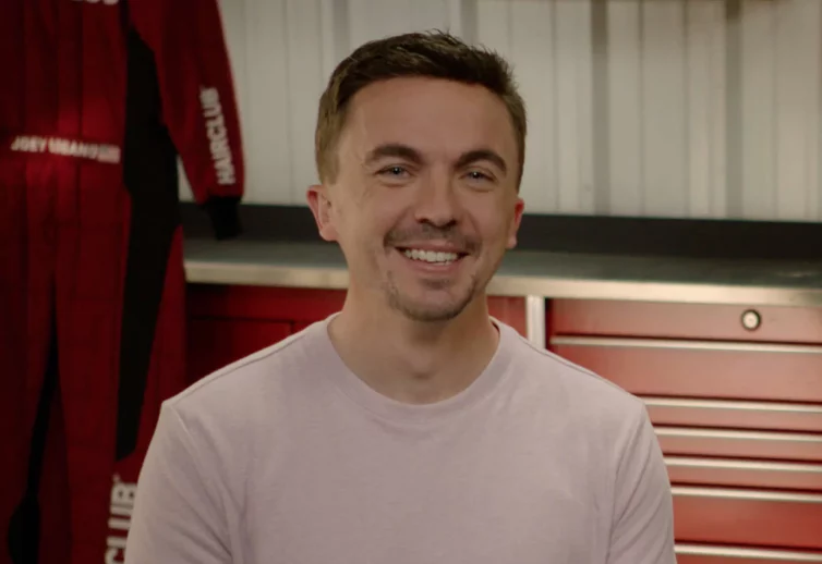 Celebrity Frankie Muniz - in the HairClub Racing garage smiling at the camera - a HairClub racing suit is hanging behind him.