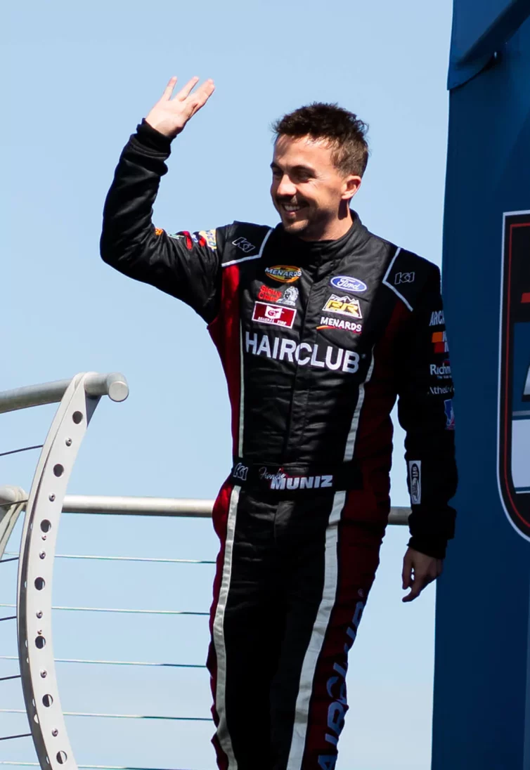 Celebrity Frankie Muniz - with great-looking hair because of HairClub Xtrands+ solution - he is outside wearing his HairClub racing suit and waving.