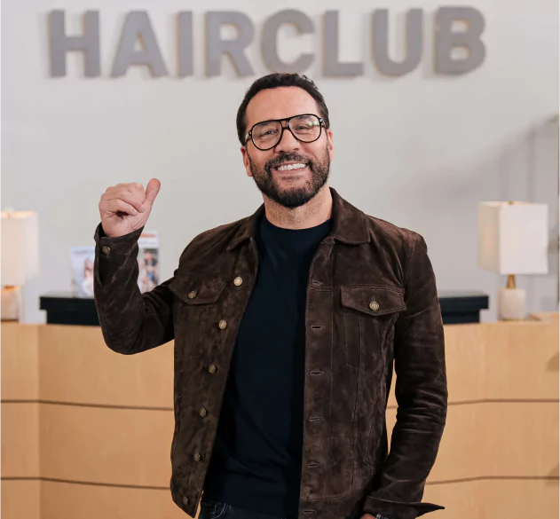 Celebrity Jeremy Piven - standing in the reception area of a HairClub Center - he is using his thumb to point to the the HairClub logo on the wall behind him.