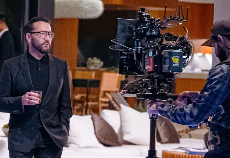 Celebrity Jeremy Piven - looks stylish in a designer black suit while holding a rocks glass and standing in front of a high-end video camera - he is in a home setting.