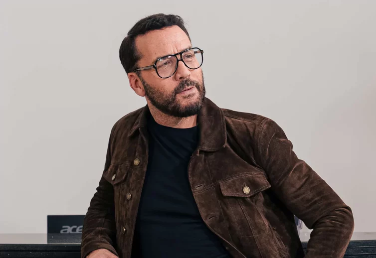 Celebrity Jeremy Piven - in a black t-shirt, a lightweight brown jacket, and black glasses - he has nice hair and is looking to the right.