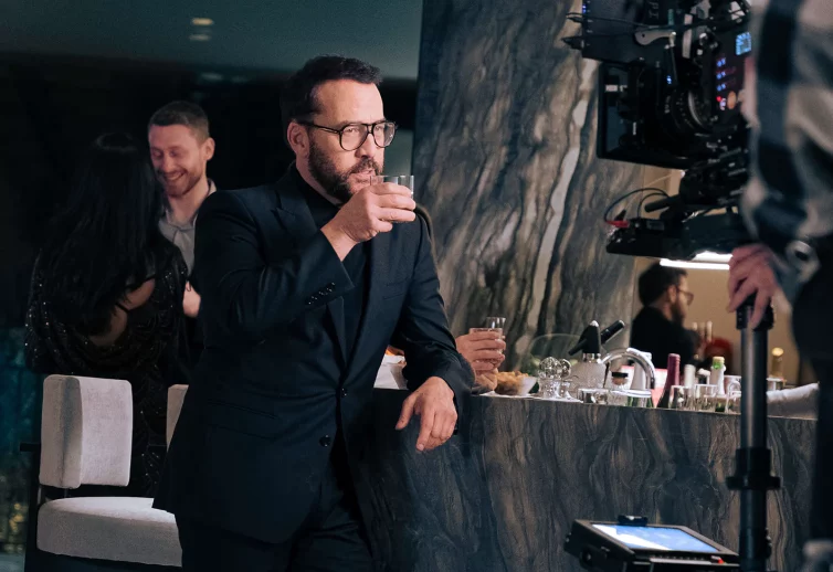 Celebrity Jeremy Piven - looks stylish in a designer black suit while holding a rocks glass near his mouth to take a sip - he is in a party setting.