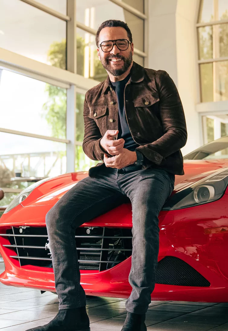 Celebrity Jeremy Piven - smiling while sitting on the hood of a red sports car.