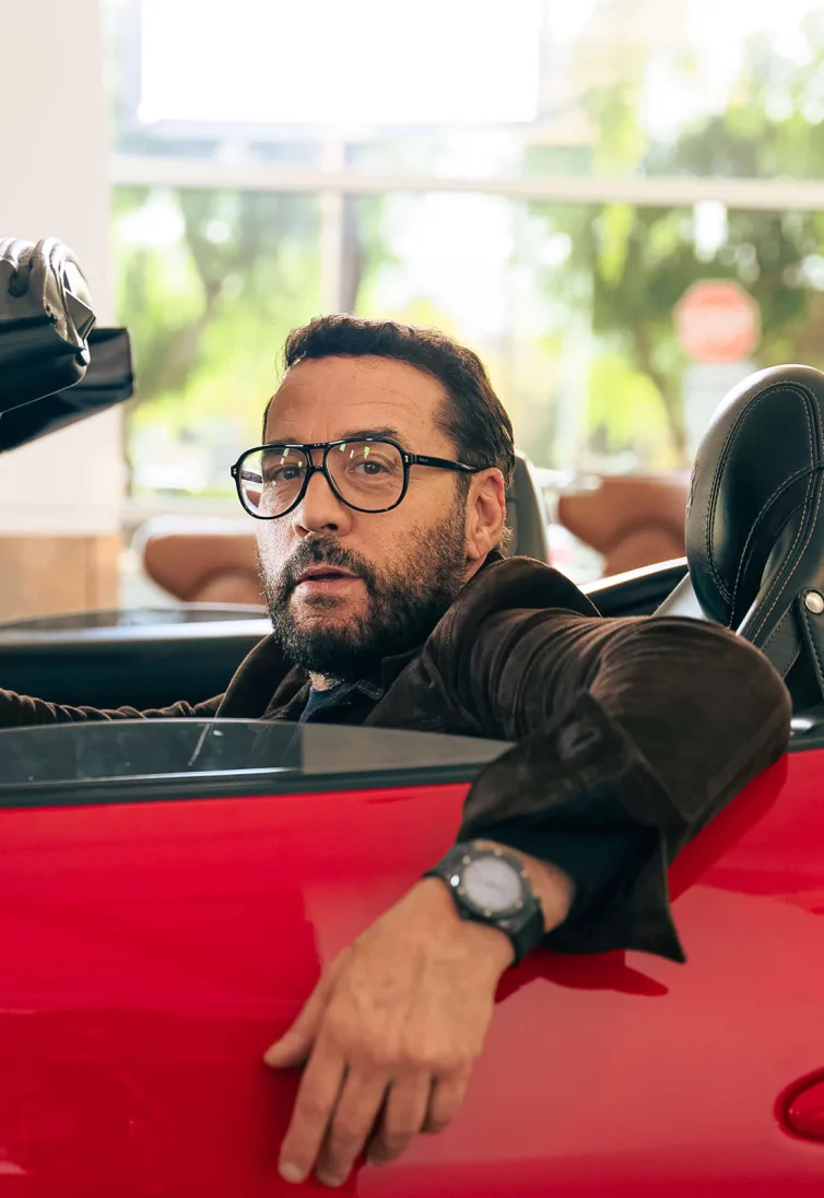 Celebrity Jeremy Piven - sitting in a red convertible car with the top down - he is looking at the camera past the rolled-down driver-side window.