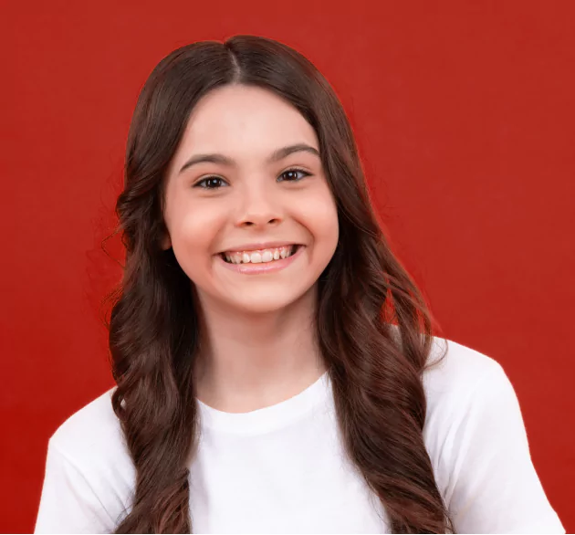 Hair Club for Kids - Learn - a portrait of a young girl in a white shirt - she has long brown hair that has been curled and has a big smile because of her HairClub hair loss solution - the background is a bold red.