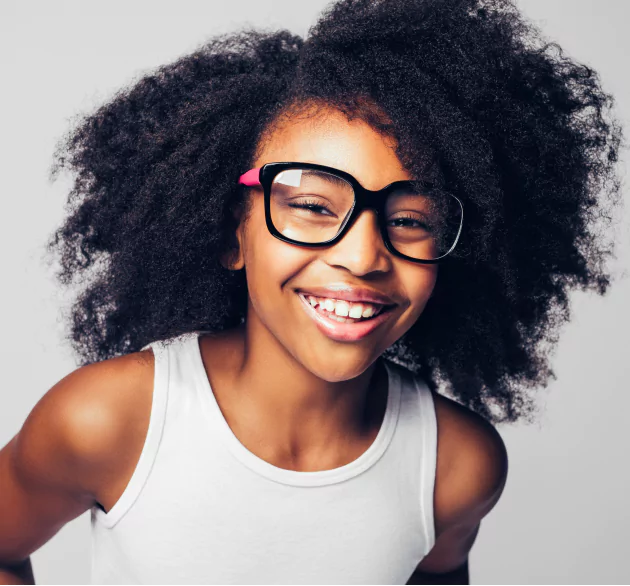 Hair Club for Kids - Learn - a portrait of a young girl with stylish, thick-rimmed glasses, wearing a white tank top - she has a big smile and a full head of thick, black, curly hair.