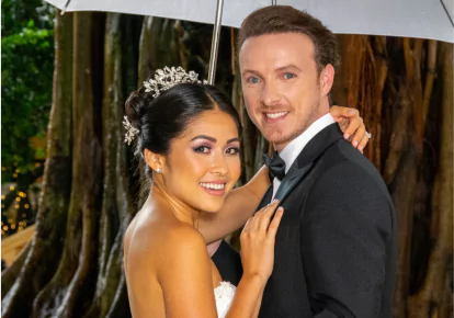 HairClub BioGraft Natural Looking Results - a bride and groom posing for a photo outside in the rain - she has her left hand around his right shoulder and her right hand on his chest - he is holding an umbrella in his right hand - their hair is perfect.
