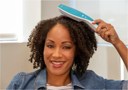 HairClub - Laser Treatment -Thicker Hair - a woman with a full head of very curly black hair uses a laser light therapy device on her head with her left hand.