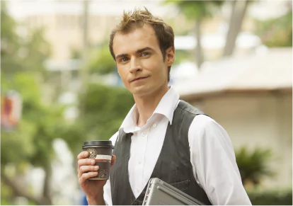 HairClub - Xtrands - Natural and Undetectable - a man with natural-looking hair wears a white dress shirt and gray vest, holding a coffee and a laptop - he is looking directly at the camera.