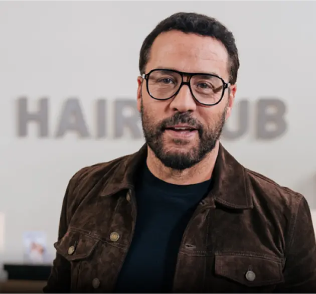 Celebrity Jeremy Piven - Testimonial - standing in front of a HairClub Center reception desk with the HairClub logo hanging on the wall behind him.